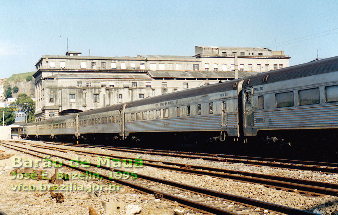 Trem de Prata visto do outro lado dos trilhos, tendo ao fundo a estação ferroviária Barão de Mauá, ex-Leopoldina, de 1926