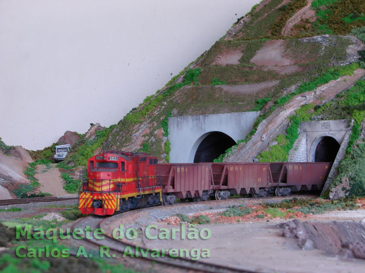 O detalhamento do cenário da maquete aumenta o realismo da locomotiva arrastando os vagões de minério sobre os trilhos em curva